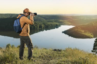 Sırt çantası ve kamera ile canlı sarı ceketli bir yürüyüşçü gün batımı dağlarının fotoğrafını çekiyor. Seyahat yaşam tarzı konsepti macera gezileri açık hava tatilleri.