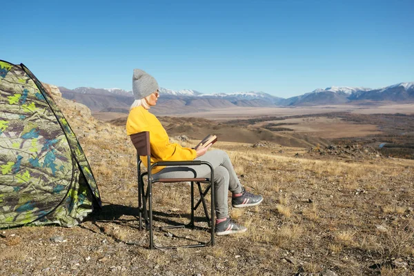 Mature woman sitting on camping chair reading a book in the autumn mountains.