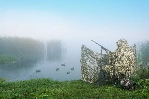 Waterfowl hunter with gun in duck hunting shelter at sunrise. Hunting with ducks decoy on lake.