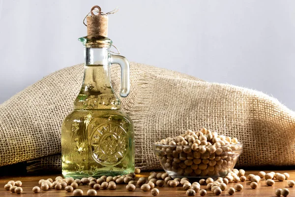 soybean seeds in a bowl with a bottle of refined soybean oil beside it on a wooden table and a jute bag in the background