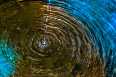 Bir damlacık, sakin bir karanlık havuzu rahatsız ederek dalga yayılmasını, enerji transferini ve doğadaki dalgalanma etkisini gösteren eş merkezli dalgalanmalar oluşturur..