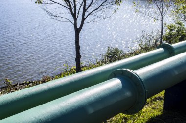 Two large green industrial pipes run parallel along a riverbank. This image represents infrastructure for water supply, sanitation, or oil and gas transportation, highlighting engineering and environmental interface.