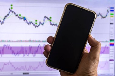 Borsa grafiğiyle ekranda akıllı bir telefon tutan bir adam, dijital çağda mobil ticareti, yatırımı ve finansal analizi sembolize ediyor..