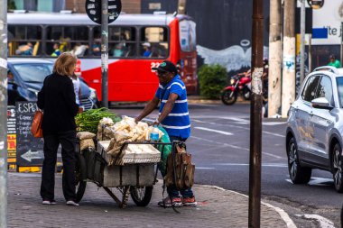Marilia, SP, Brezilya, 19 Ağustos 2025. Şehir merkezindeki işlek bir caddede, bir sokak satıcısı müşterisine taze ürünlerini sunuyor. Satıcının arabası, içi manyok, fıstık ve diğer şeylerle dolu.