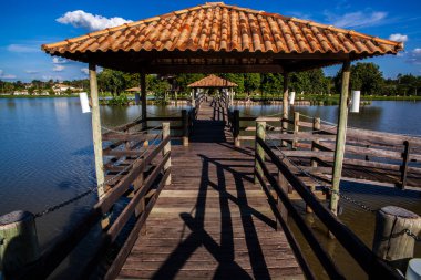 Brezilya 'da güneşli bir günde sakin bir göle uzanan fayanslı çatılı ahşap iskele. Yeşil doğa ve açık mavi gökyüzü ile çevrili boş bir yol, turizm ve rahatlama için mükemmel..