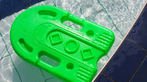 Canlı yeşil bir kickboard bir yüzme havuzunun kenarındaki dalgalı suyun üzerinde duruyor. Parlak renk, havuz sınırının beyaz ve koyu mavi karolarına karşı güçlü bir karşıtlık sağlar.