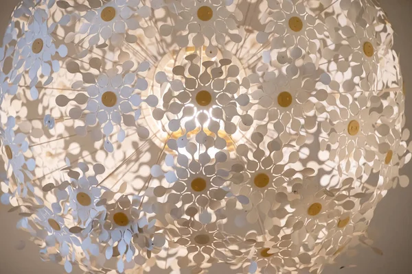 Sarı merkezli sayısız beyaz plastik çiçek yaprağından oluşan geniş, küresel bir avizeye bakarak detaylı bir yakın çekim görüntüsü. Merkezi ışık kaynağı dramatik gölgeler yaratır ve parlar.