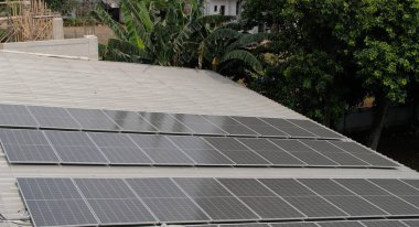 Gri kıvrımlı metal çatıya yerleştirilmiş büyük bir fotovoltaik güneş paneli dizisi. Paneller, temiz, sürdürülebilir ve yenilenebilir enerji teknolojisini sembolize ederek güneş ışığını yakalıyor.