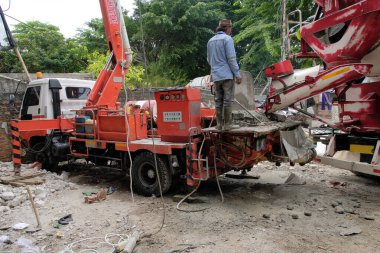 Kauçuk çizmeli inşaat işçileri yeni döşenmiş zemine taze, ıslak beton dökmek için geniş esnek bir pompa borusu kullanıyorlar. Sahne endüstriyel yapı, Jakarta, Aralık 2025 'i vurgular