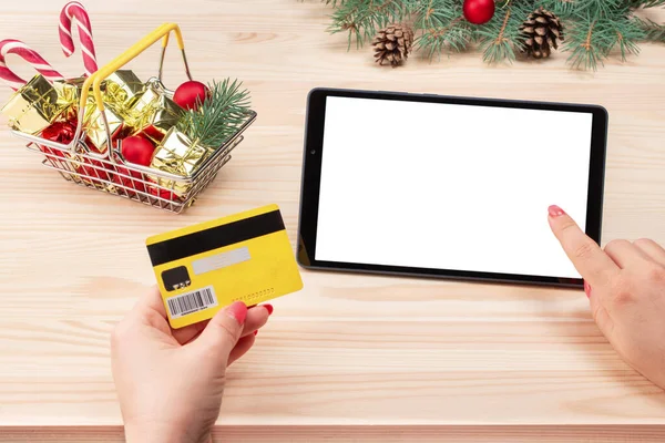 women's hands hold a credit card and mockup tablet on background of basket with Christmas gifts. Christmas online shopping with tablet device. Christmas Wooden desk on background