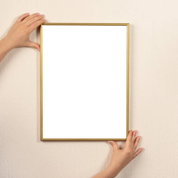 Woman hanging a frame mockup on a wall. young woman\'s hands hold a gold frame on a light wall background. Certificate, diploma, picture, gratitude white frame mockup. square photo