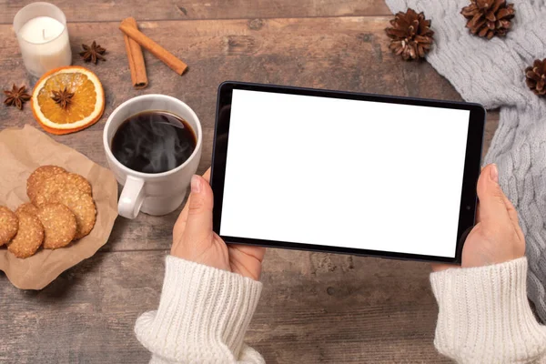 Mockup tablet pc. Woman\'s hands holding black tablet pc with blank white screen while drinking coffee on wooden table in cafe. winter Christmas time