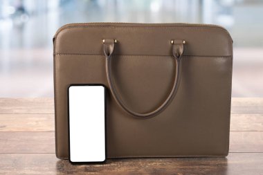 Cep telefonu modeli ve ahşap bir masanın üzerindeki deri bir iş çantası bulanık bir ofis geçmişine karşı. Boş ekranlı cep telefonu