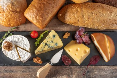 Siyah taş tahtaya farklı peynirler serilmiş. Mavi Dorblu peyniri, füme Suluguni peyniri, peynir, Maasdam peyniri, çedar peyniri, cevizli parmesan peyniri, üzüm, domates ve ekmek. en üstteki görünüm