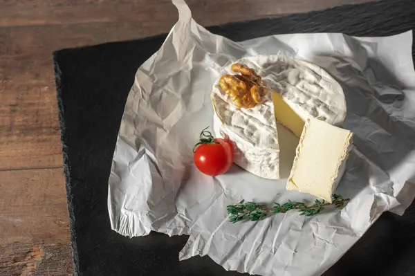 Delicious brie cheese, walnuts and tomato on paper on slate black background. Brie type of cheese. Camembert. Fresh Brie cheese and a slice on stone board. Italian, French cheese.