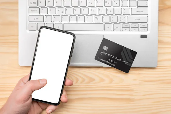 Online paying shopping, electronic payment with credit card, cell phone mockup, laptop over wooden table. man hand holding smart mobile phone, using credit card for online shopping with laptop