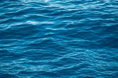 Parlak güneş ışığı yansımalı mavi su yüzeyi, okyanusta su veya deniz arka planında yakın çekim. Dalgalı ve köpüklü mavi deniz dokusu. Mavi deniz suyu yüzeyi