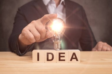 Elinde ampul, fikir, yenilik ve ilham konsepti olan iş adamının eli. Üzerinde IDEA metni olan tahta küpler. Yenilik ve yaratıcılıkla yeni bir fikir oluştur