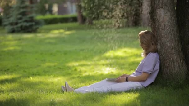 Junge schöne Frau arbeitet an Tablet-Computer sitzt auf Gras im Park — Stockvideo