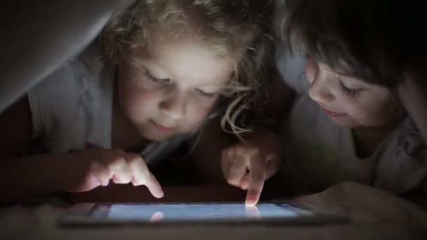Две маленькие девочки-сестры рисуют на планшетном компьютере, прячутся под одеялом — стоковое видео