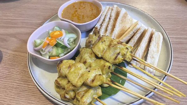 Kremalı fıstık sosu, salatalık çeşnisi ve ahşap bir masada dilimlenmiş ekmek ile servis edilen Tayland Pork satay şişleri. Izgara şişte altın bir zerdeçal salamurası var.