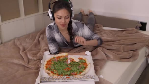 Młoda kobieta leżąca na łóżku je pizzę. — Wideo stockowe