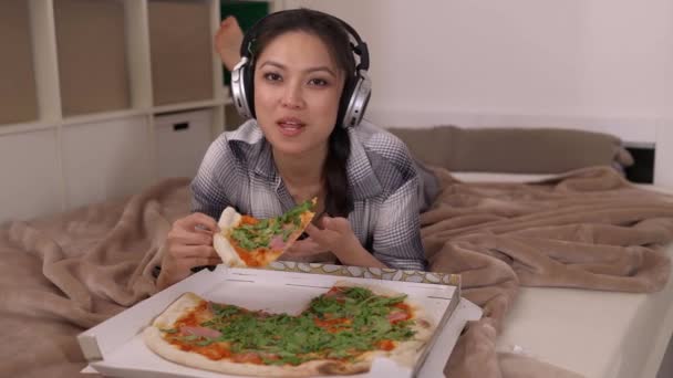Молодая женщина лежит на кровати и ест пиццу. — стоковое видео