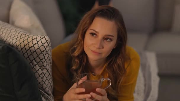 Junge hübsche Frau trinkt eine Tasse Tee, während sie auf der Couch liegt — Stockvideo