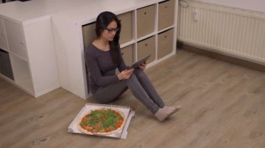 Genç bir kadın tablet bilgisayarıyla yerde oturuyor ve pizza yiyor.