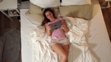 Genç seksi kadın yatağında tablet bilgisayarıyla uzanıyor.
