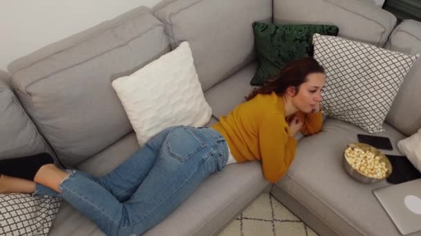 Молода жінка лежить на дивані і дивиться телевізор — стокове відео
