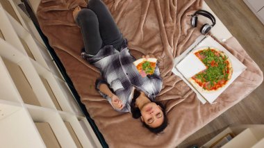 Genç ve güzel bir kadın yatağa uzanıp pizza yiyor.