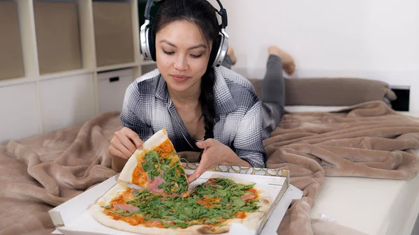 Młoda kobieta leżąca na łóżku je pizzę. — Zdjęcie stockowe