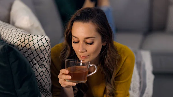 Giovane bella donna beve una tazza di tè mentre sdraiato sul divano — Foto Stock