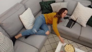 Genç bir kadın dizüstü bilgisayarıyla kanepede uzanıyor ve patlamış mısır yiyor.