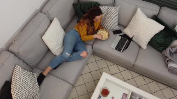 Młoda kobieta leży na kanapie z laptopem i je popcorn — Wideo stockowe