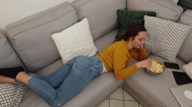 Genç bir kadın kanepede uzanıyor ve televizyon izliyor.