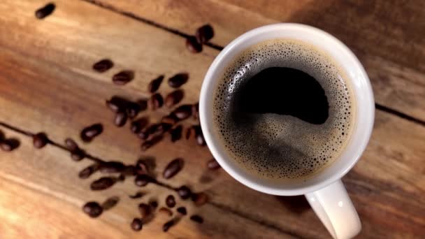 Свежесваренная чашка кофе на деревянном столе - кофейные зерна падают в замедленной съемке — стоковое видео