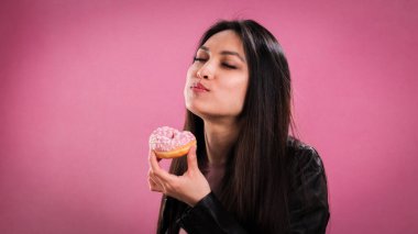 Genç bir kadın pembe arka planda donut yiyor.