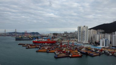 BUSAN, Güney Kore - 9 Ekim 2025 Busan limanı kargo gemileri, mavnalar, şehir binaları ve bulutlu bir gökyüzü altında dağlarla dolup taşıyor