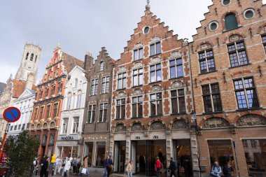 BRUGES, NETHERLANDS - 15 Eylül 2025 - Belçika 'nın Bruges şehrinde yürüyen ve alışveriş yapan insanlar, ünlü çan kulesi renkli tuğla binaların üzerinde yükseliyor