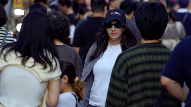 BUSAN, Güney Kore - 9 EKİM 2025 - Kore şapkası takan ve güneş gözlüğü takan kadın şehir sokaklarında kalabalığın arasında yürüyor