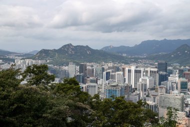 SEOUL, Güney Kore - 11 Ekim 2025 - Seul 'ün yeşil ağaçlar ve bulutlu bir gökyüzünün altındaki uzak dağlarla çevrili modern binaları ile şehir silueti