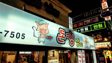 BUSAN, Güney Kore - 10 EKİM 2025 - Parlak neon tabelalar, Kore yemeklerinin reklamını yapan bir domuzun olduğu sokak sahnesini aydınlatır