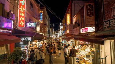 BUSAN, GÜNEY KOREA - 10 EKİM 2025 - İnsanlar parlayan neon ışıkları ve sokak yiyecek tezgahlarıyla çevrili Busan gece pazarının canlı atmosferinin tadını çıkarıyorlar