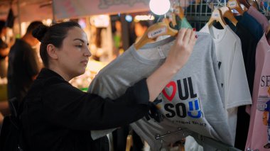 SEOUL, GÜNEY KORE - 11 Ekimde. 2025 - Seul 'deki hareketli bir açık hava pazarında kadın giysi raflarına göz atıyor, hediyelik bir tişört seçiyor.