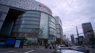 BUSAN, GÜNEY KORE 10 EKİM. 2025 - Bulutlu bir gökyüzünün altında büyük Lotte AVM binası olan bir caddede insanlar ve arabalar hareket ediyor