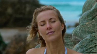 Şık bikinili canlı bir kadın okyanusun kıyısındaki bir kayanın üzerinde oturur, yumuşak dalgaların ve ılık güneşin tadını çıkarır.