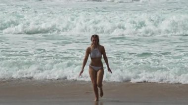 Bir kadın kumsalda güzel bir günün keyfini çıkarıyor, parlak, berrak bir gökyüzünün altındaki dalgalara doğru yürüyor.