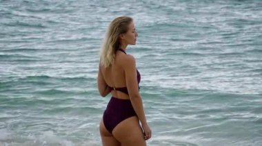 Şık bikinili bir kadın okyanusun kenarında zarif bir şekilde duruyor, sakin ve sakin sahil atmosferinin tadını çıkarıyor.